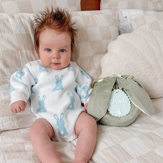 Baby wearing a white onesie with blue bunny patterns, sitting on a couch next to a green bag.