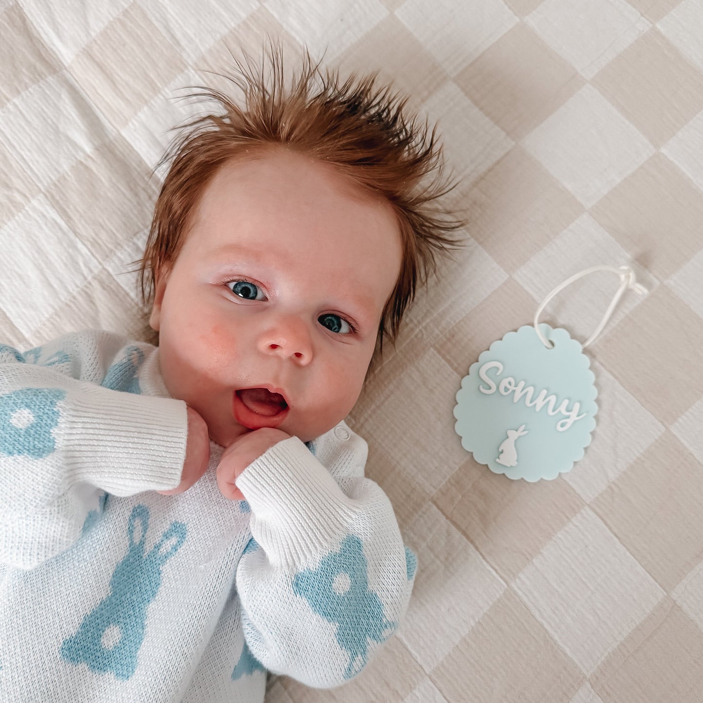 Newborn baby wearing a white sweater with blue patterns, lying on a checkered blanket.